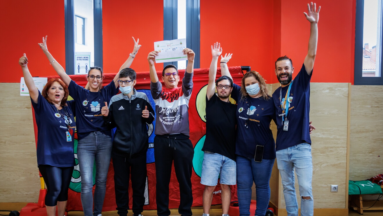 En las instalaciones de Down Madrid en Madrid aparecen 7 personas, cuatro trabajadores de Amazon y tres participantes de la fundación con los brazos levantados. Los trabajadores de Amazon van con la misma camiseta azul y el badge colgado y los de Down Madrid con deportivas y ropa cómoda. El fondo es de color rojo y el suelo de parque de pista de baloncesto cubierta.