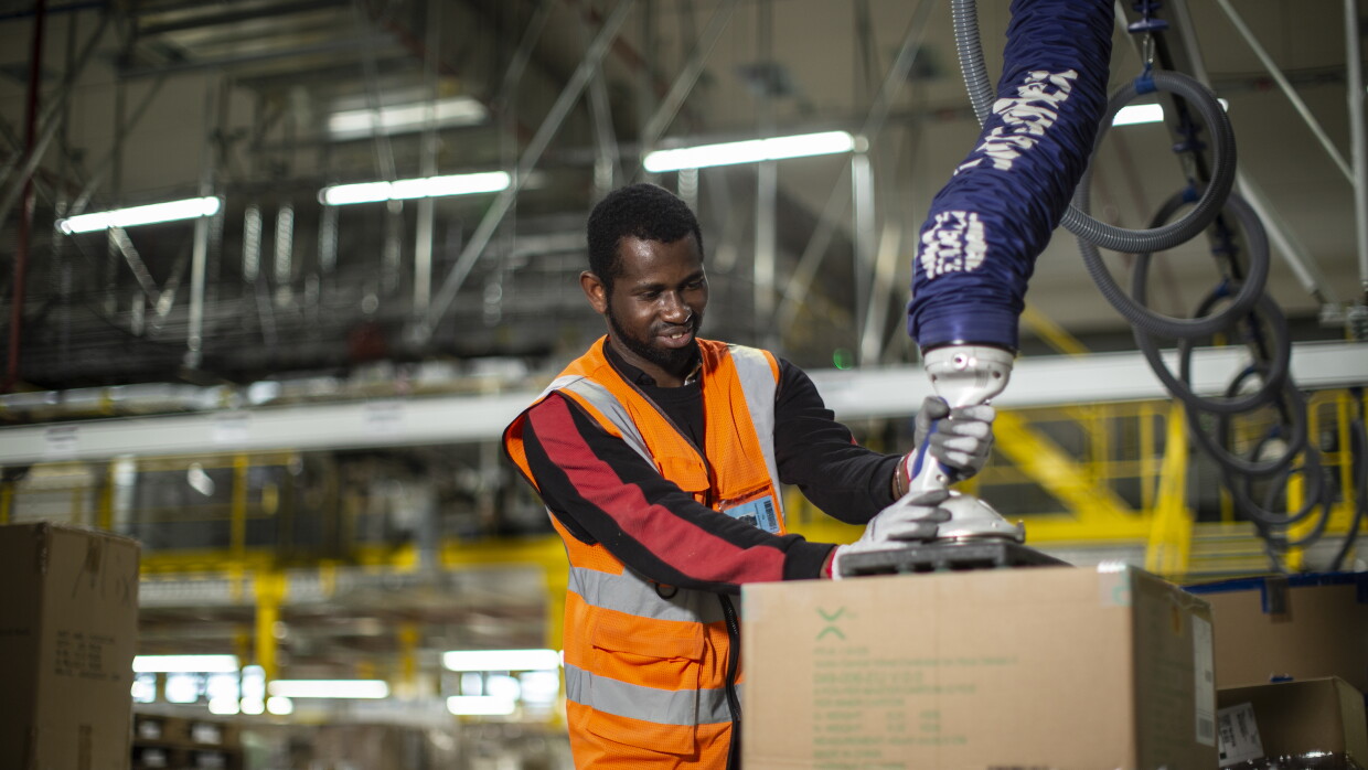Un dipendente Amazon lavora con un macchinario in un magazzino. Indossa un gilet catarinfrangente e dei guanti da lavoro. 