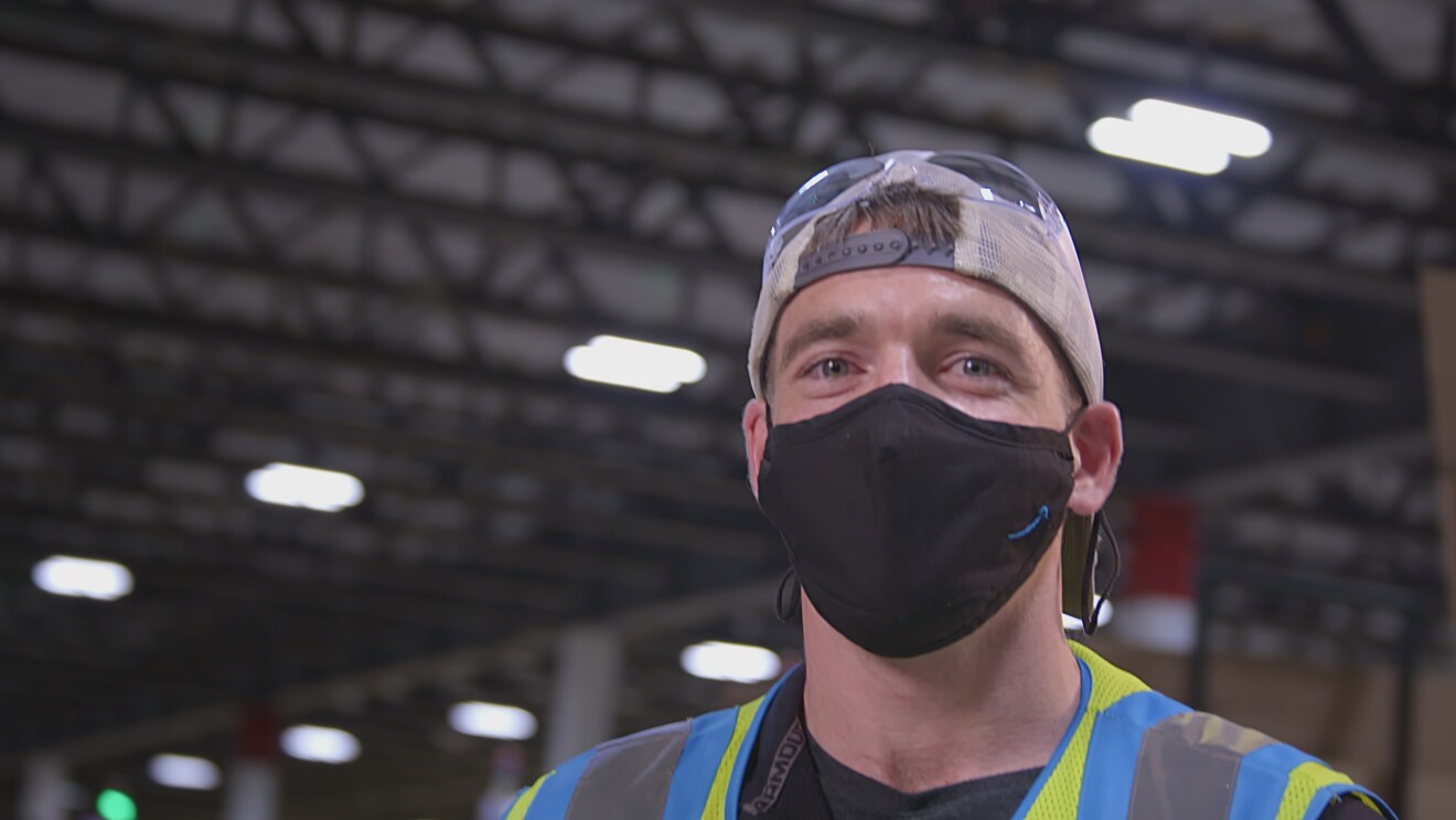 An Amazon employee poses from inside an Amazon Fulfillment Center wearing a backwards cap, black mask, and high visibility vest.