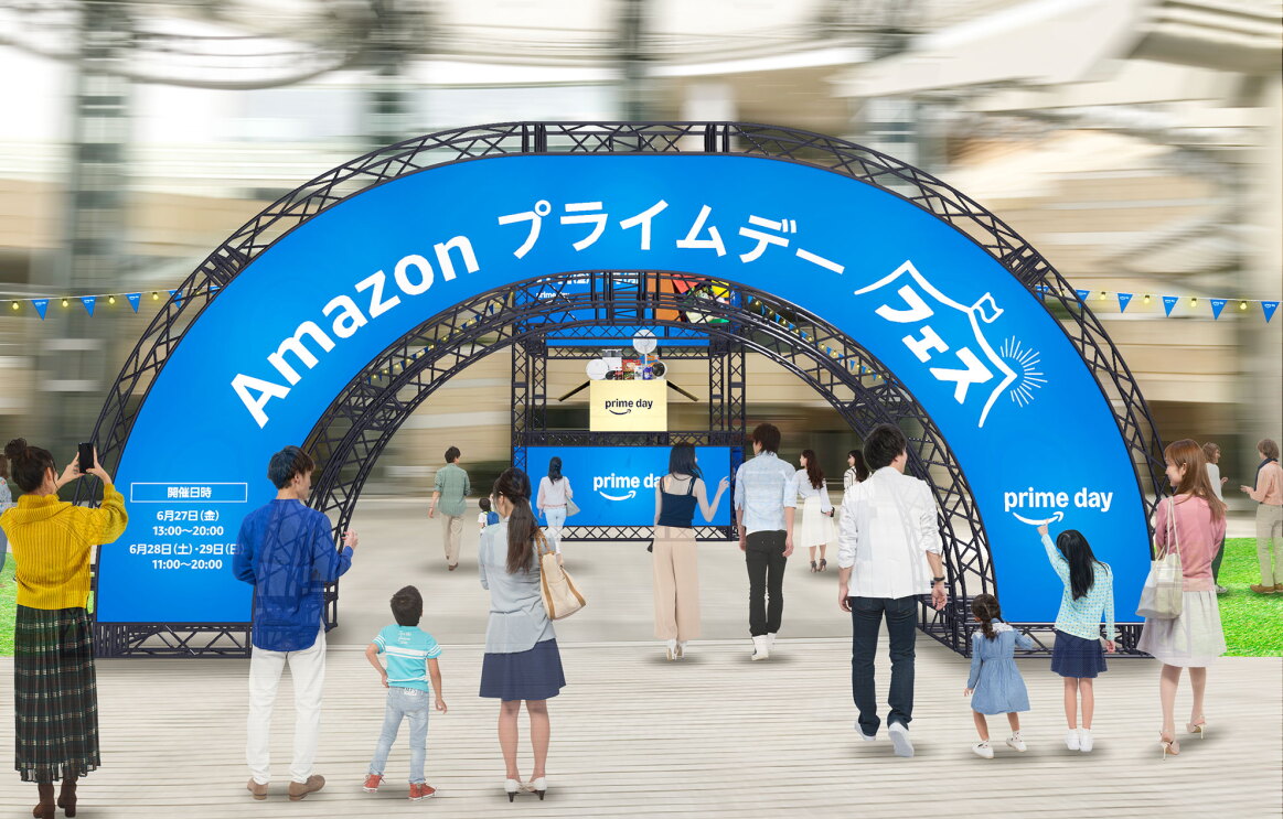 イベント会場に設置されたAmazon プライムデーの青いアーチ型バナー。大きく白字で「Amazonプライムデーフェス」と書かれている。アーチをくぐって入場する人、アーチを指さす家族連れ、アーチの写真を撮る人などがいる。