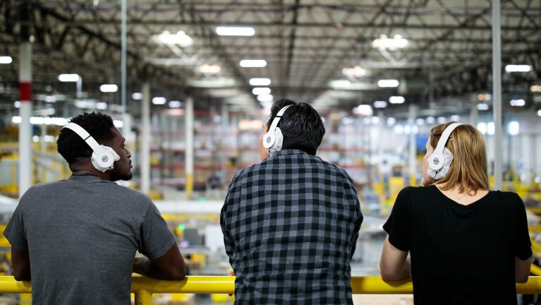 広大な物流倉庫の内部。高い天井と無数の照明が奥まで続く。手前には白いヘッドフォンを着けた私服の人物が3人いて、黄色い手すりに寄りかかって倉庫を眺めている。