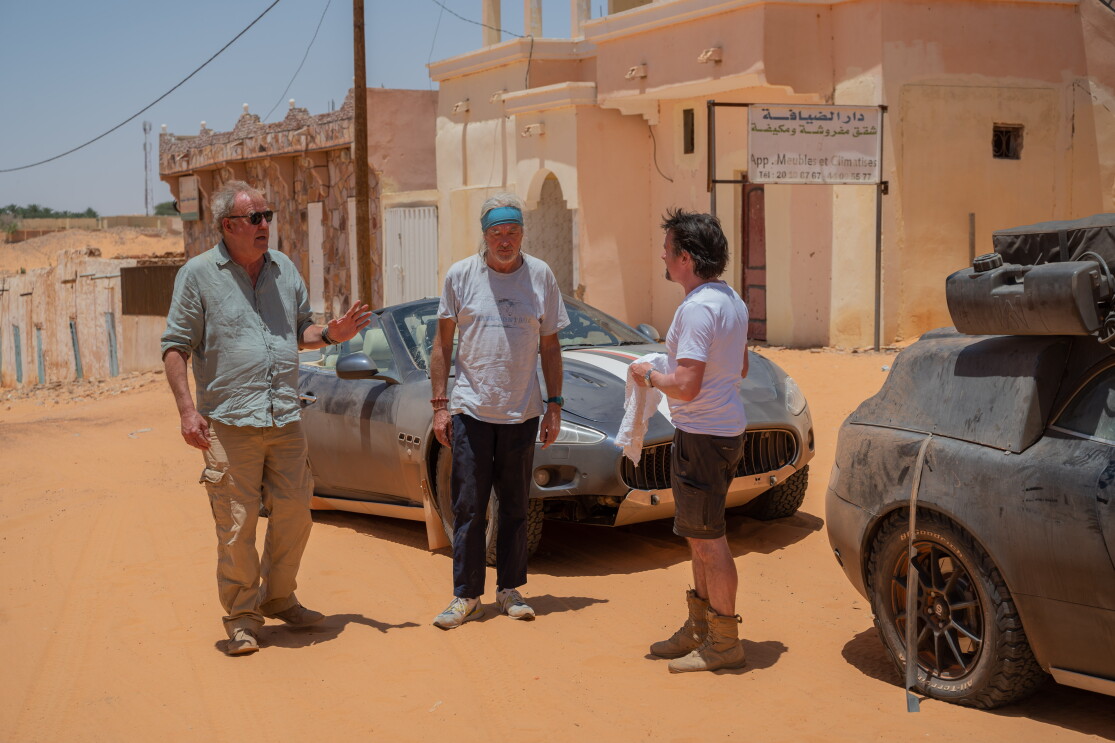 Three older men stand around a sports car 
