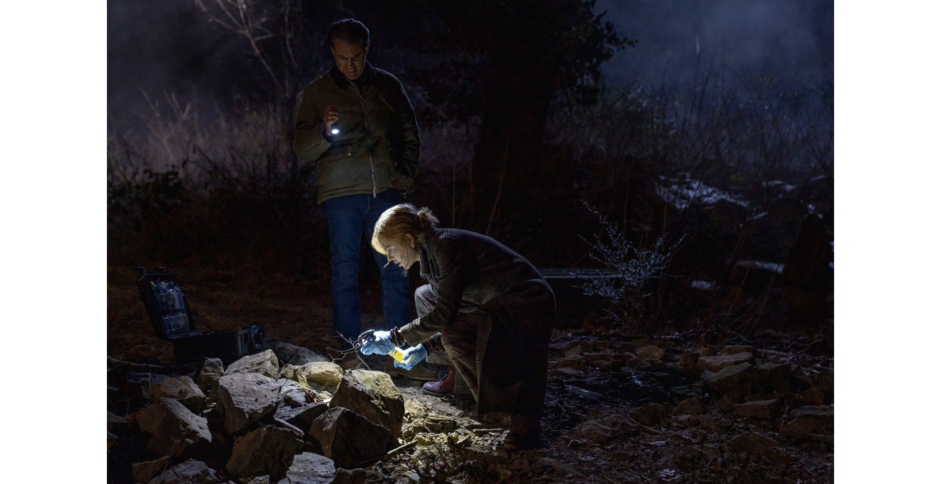 夜間の暗い屋外で、懐中電灯を持った2人が岩の多い地面を照らして調べている。手間の人物は青い手袋を着用、かがんでいる。