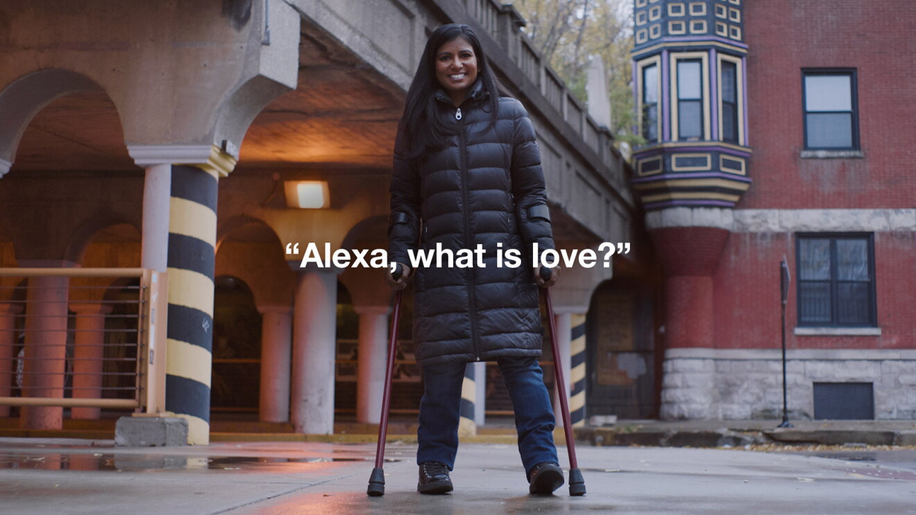 An image of a woman smiling for a photo while standing in an entryway while using crutches. Words that say "Alexa, what is love?" overlay the image.