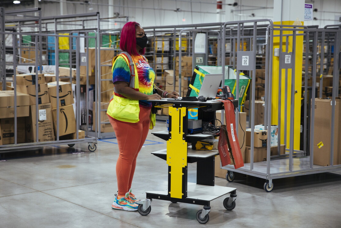 Auhzha Wright stands at her computer workstation in front of boxes and rolling carts inside Amazon's Fulfillment Center