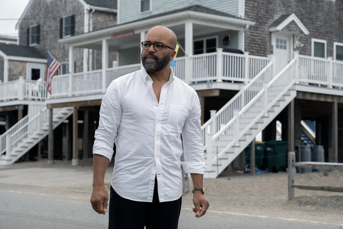 A bald man in a white shirt and black pants standing in front of a house.