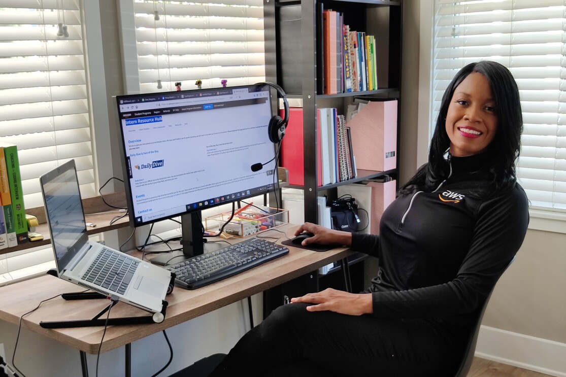 A woman sits at a desk with a laptop and a computer monitor.