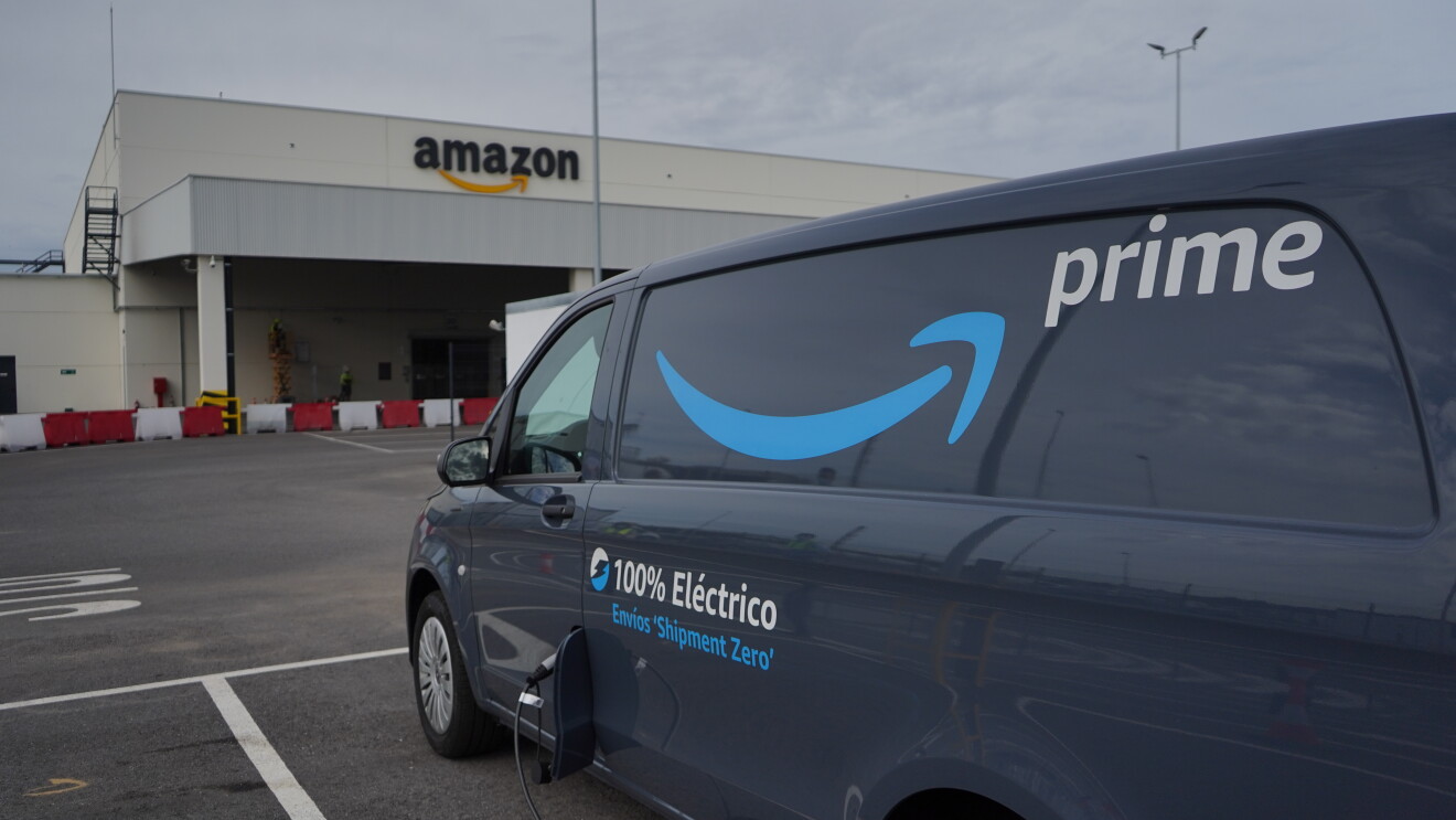 Una Mercedes Benz-Vans en un aparcamiento. Se ve el logo Prime de Amazon y el texto en la puerta 100% Eléctrico Envíos 'Shipment Zero'. En primer plano se ve un cargados de batería que está subministrando electricidad a la furgoneta de al lado que solo se ve un poco su parte trasera. 