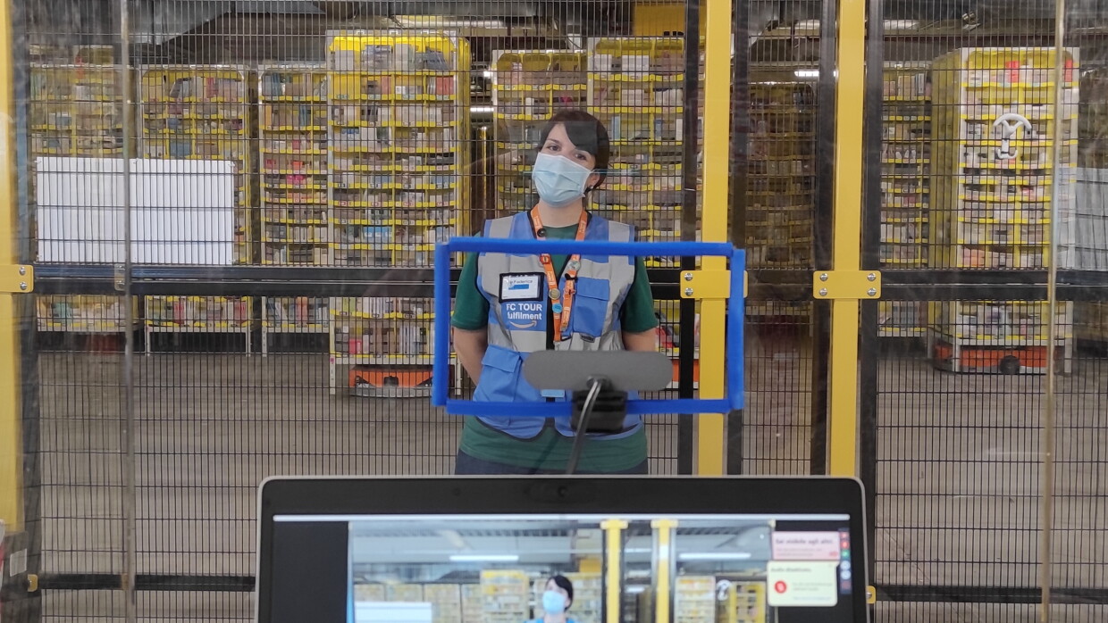 In primo piano un tablet con la fotocamera accesa. Sullo schermo si vede una ragazza all'interno di un magazzino Amazon. La ragazza è posizionata di fronte al tablet, indossa una mascherina chirurgica e un giubbino catarinfrangente blu con la scritta FC Tour Ambassador. Alle sue spalle, una griglia la separa dall'area robotica del magazzino, in cui si vedono le colonne di prodotti trasportate dai robottini. 