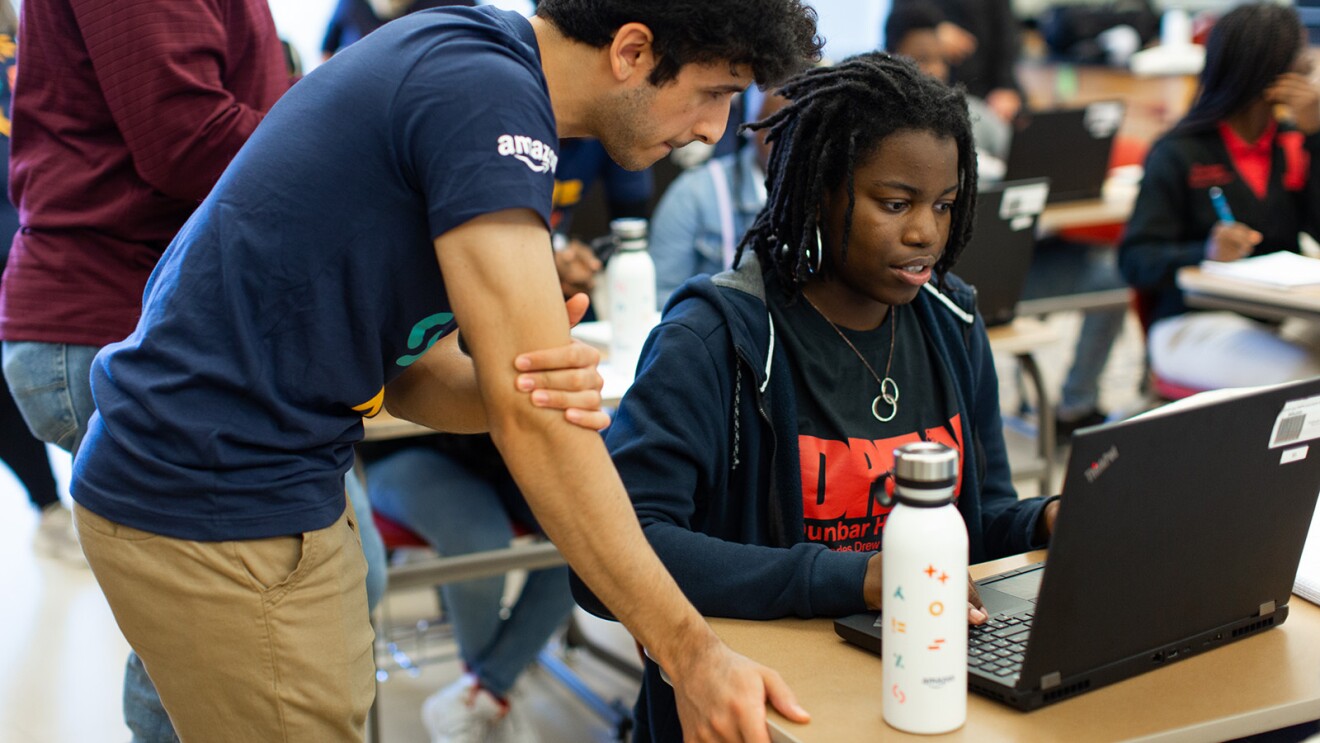 A photo of an instructor assisting a student who is sitting at a desk, working on a laptop device.