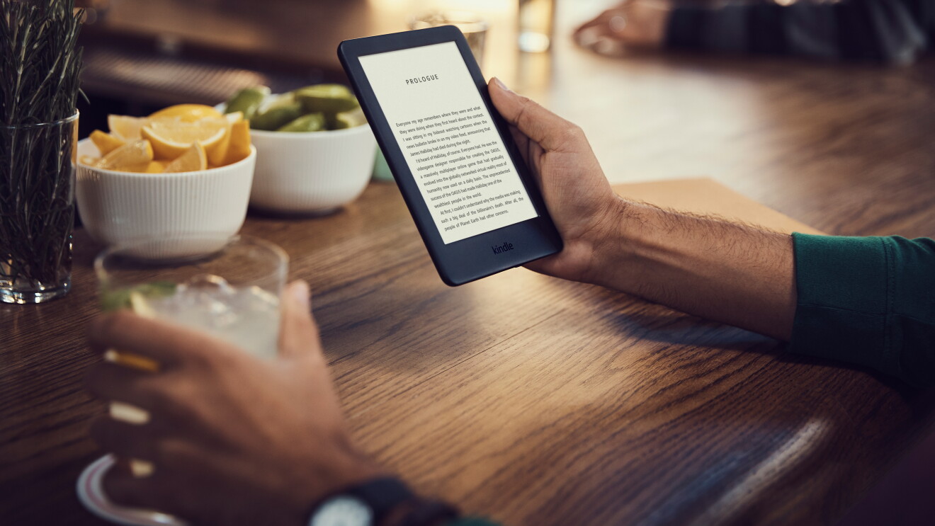 A man holds a Kindle device in his right hand, reading the prologue of a book. He holds a cocktail in the right hand, in a bar setting. 