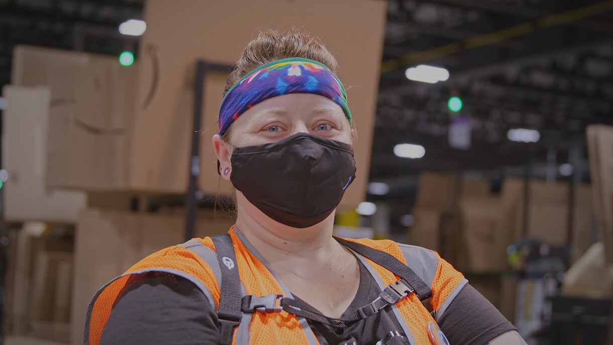 An Amazon associate wearing a mask, a high visibility vest, and a tie-died headband poses in front of a stack of flat-packed cardboard boxes in an Amazon fulfillment center