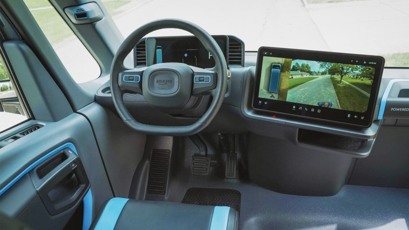 The inside driver side of the an Amazon Rivian delivery vehicle.  There is a big screen with a camera showing the outside of the vehicle.