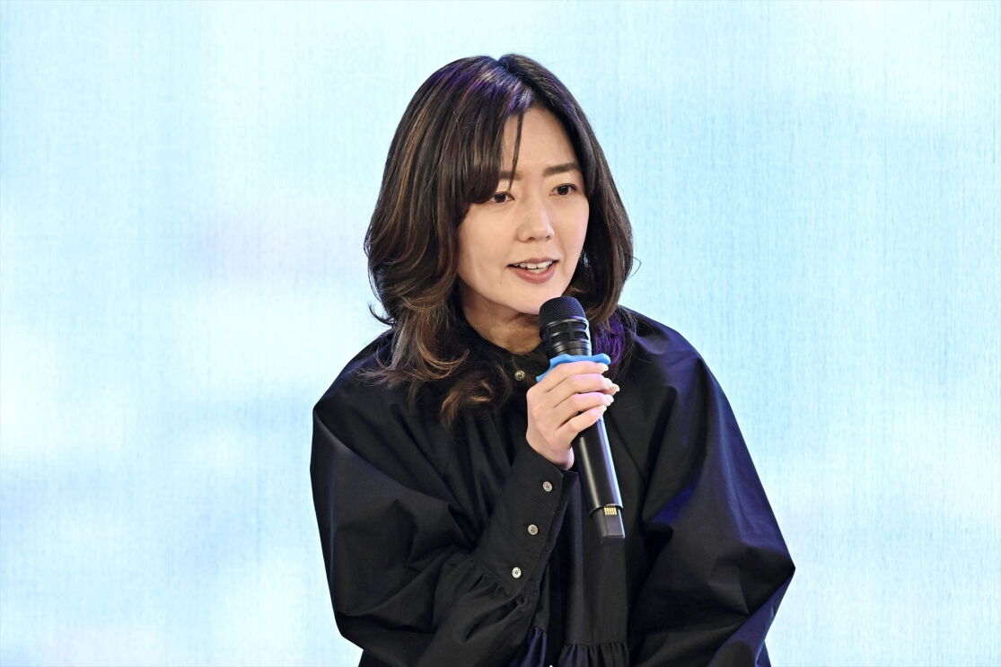 黒いロングスリーブを着ている女性がマイクで話している。川本さん。