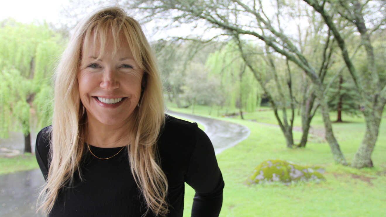 A smiling woman with blond hair wearing a black top in a park next to a cycling path. 