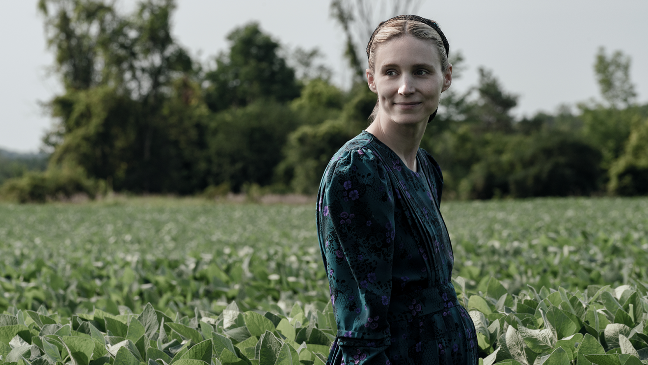 An image of a woman standing in a field, looking away and smiling. 