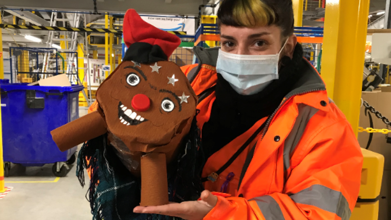 Adorno navideño de BCN8.  Una asociada con mascarilla, jersey y chaleco naranja sujeta un tió (tronco navideño típico catalán) que está hecho de cartón y tiene barretina (gorro típico catalán) y manta. 