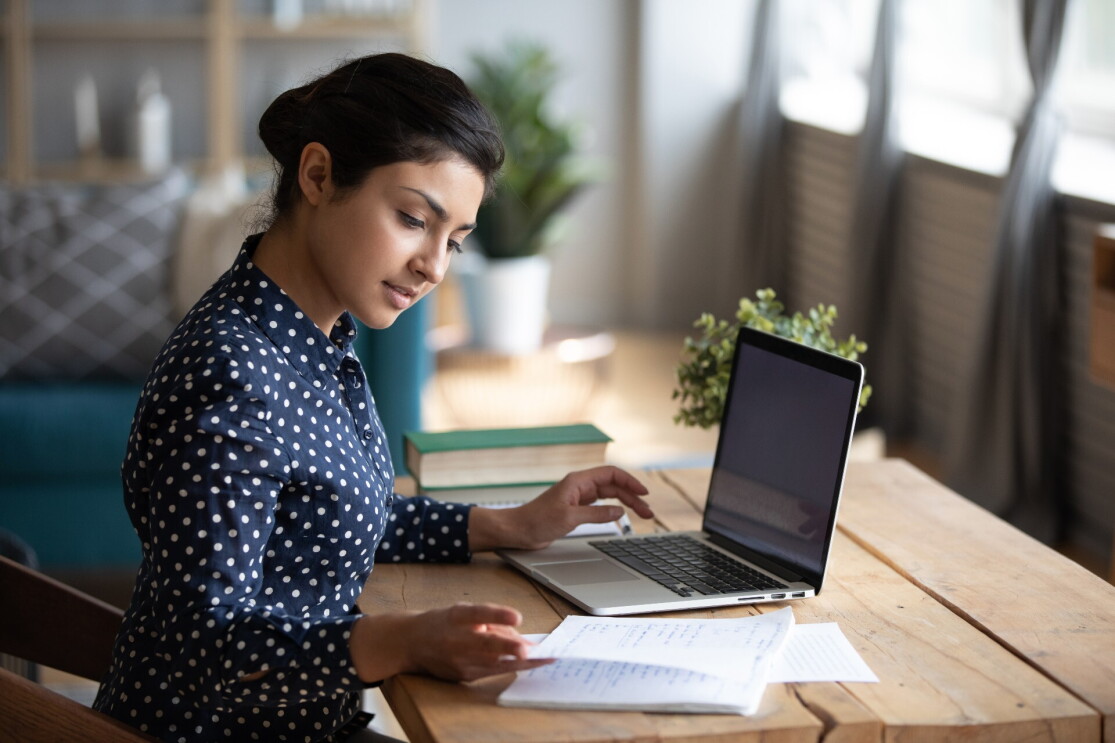 A woman wearing a polka-dotted blouse studies at her desk with her laptop and notes in front of her.
