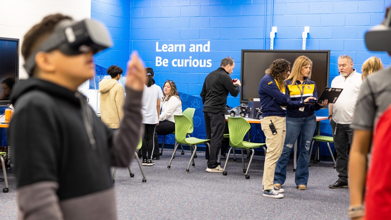 A photo of students visiting the AWS Think Big Space in at J. Lupton Simpson Middle School in Leesburg, Virginia. There is a student in the forefront of the photo trying on virtual reality goggles, and students talking with several teachers in the background.