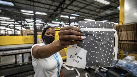 A woman wearing a face mask working at an Amazon fulfillment center holds up a small gift bag with a tag that says "A gift for you" printed on it. 