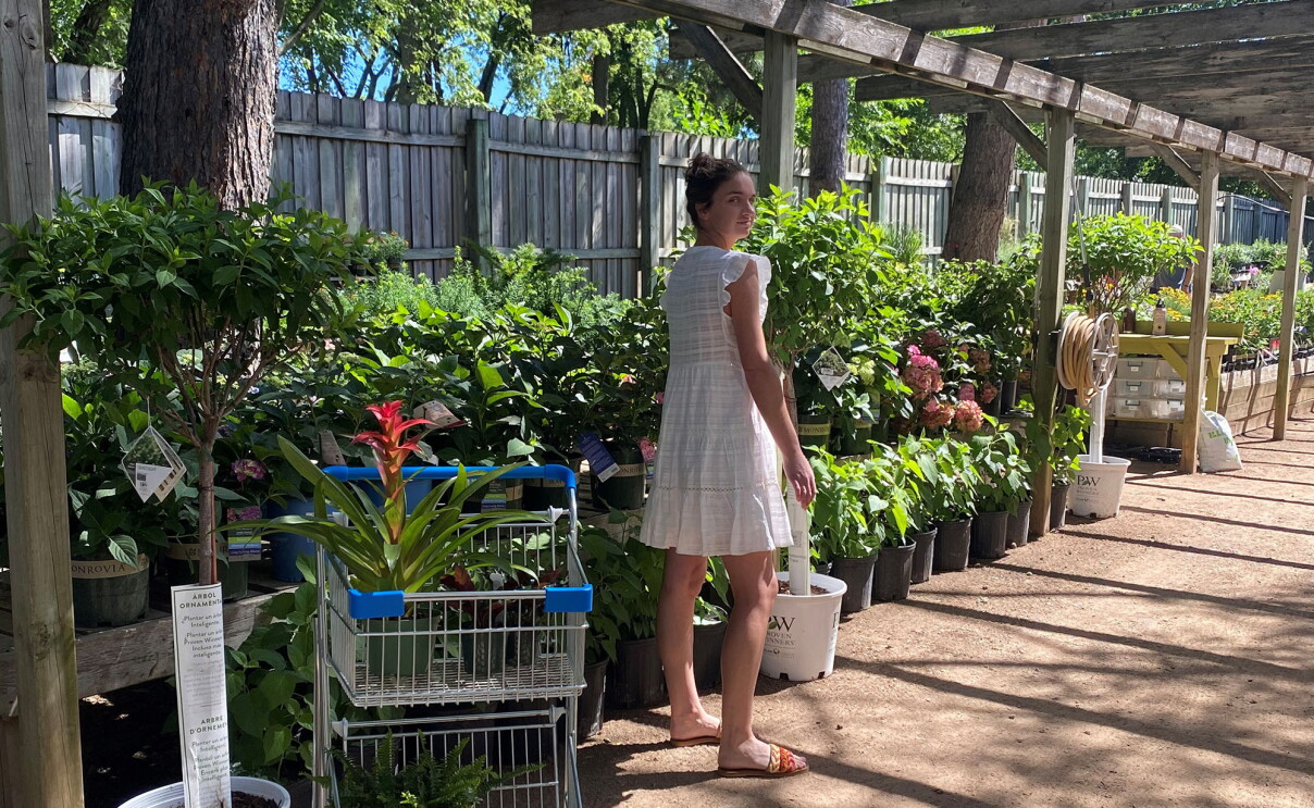 An image of a woman standing in front of a row of plants at a garden store. There is a shopping cart in front of her with a plant in it. 