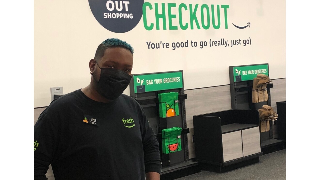 Fred wears an Amazon Fresh black long-sleeved shirt and a face mask. He poses in front of the Amazon Store where customers can bag their own groceries. The wall above the bags reads, "You're good to go (really, just go)"