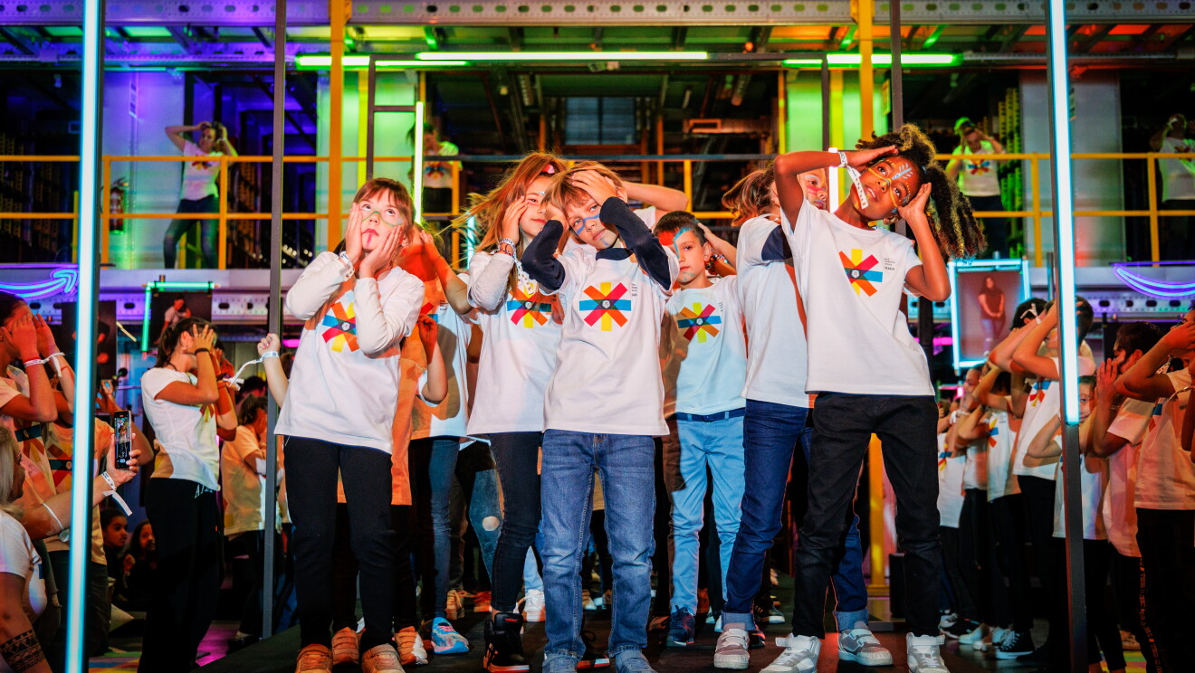 Dentro de un centro logístico, con las estanterías de fondo y las barandillas de color amarillo, aparece una pasarela de color negro ambientada con luces de neón. Un gupo de niños con jeans y camisetas blancas del evento con pinceldas de colores están realizando una correografía con las manos en la cabeza.