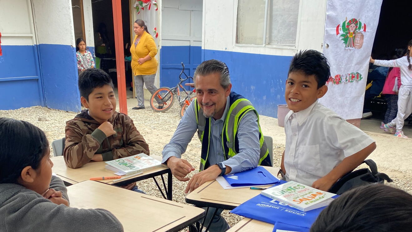 En un patio de colegio, un voluntario de Amazon con cuatro niños alrededor sentados en pupitres. 