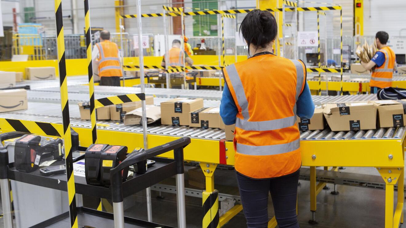 En un centro logístico aparecen cuatro asociados de espaldas trabajando en una cadena  de distribución de paquetes. Las pasarelas son de color amarillo y los asociados están colocados de espaldas con chalecos naranja. En primer plano está una asociada de espaldas manipulando cajas de Amazon. Tiene el pelo negro, recogido en una cola, con un chaleco de color naranja, una sudadera de color azul y unos pantalones jeans. 