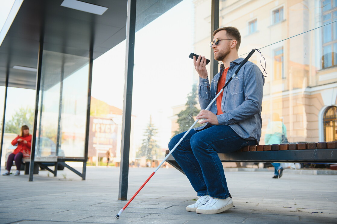 A blind man sitting on a bench speaking into a phone. 