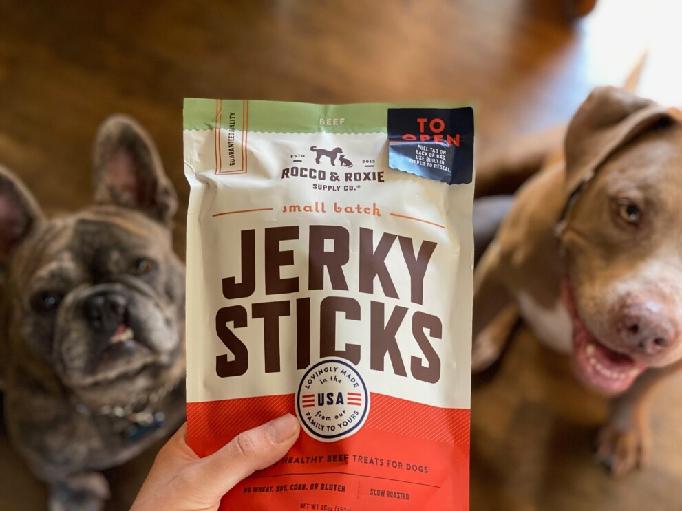 Two dogs stare up at a person holding a package of jerky sticks.