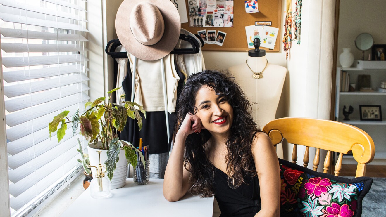 An image of a woman smiling for a photo while resting her head on her hand. She is at a desk and there is a bulletin board with various items including a wide-brim hat and photos behind her.