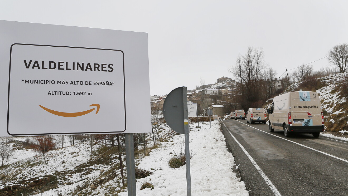 Cartel en la carretera que anuncia la entrada al pueblo. El cartel lleva el símbolo de Amazon y la inscripción: Municipio más alto de España, 1692 m. Al lado derecho hay 4 furgonetas decoradas como si fueran paquetes de Amazon y con la etiqueta deliveringsmiles. 
