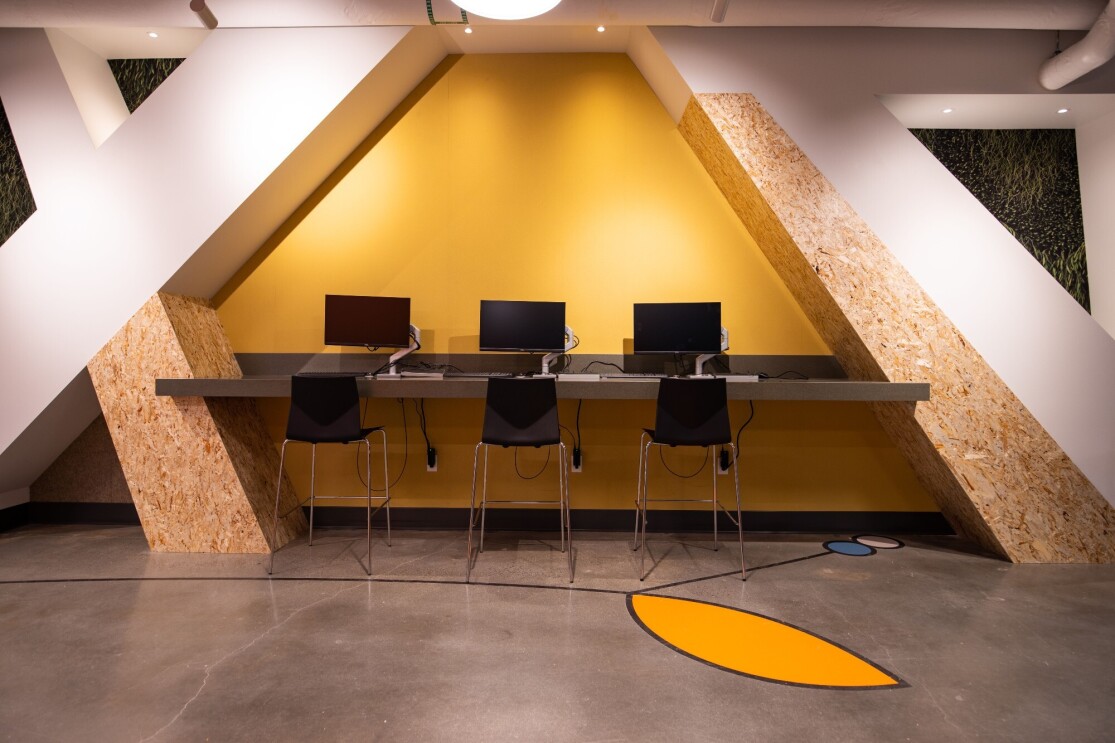 An image of a row of computers on a counter with tall stools where people can work on the computers.