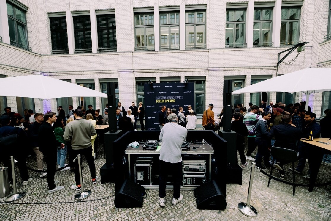 Im Innenhof des neuen Amazon Büros in Berlin stehen Gäste zusammen und unterhalten sich. Ein DJ legt Musik auf.