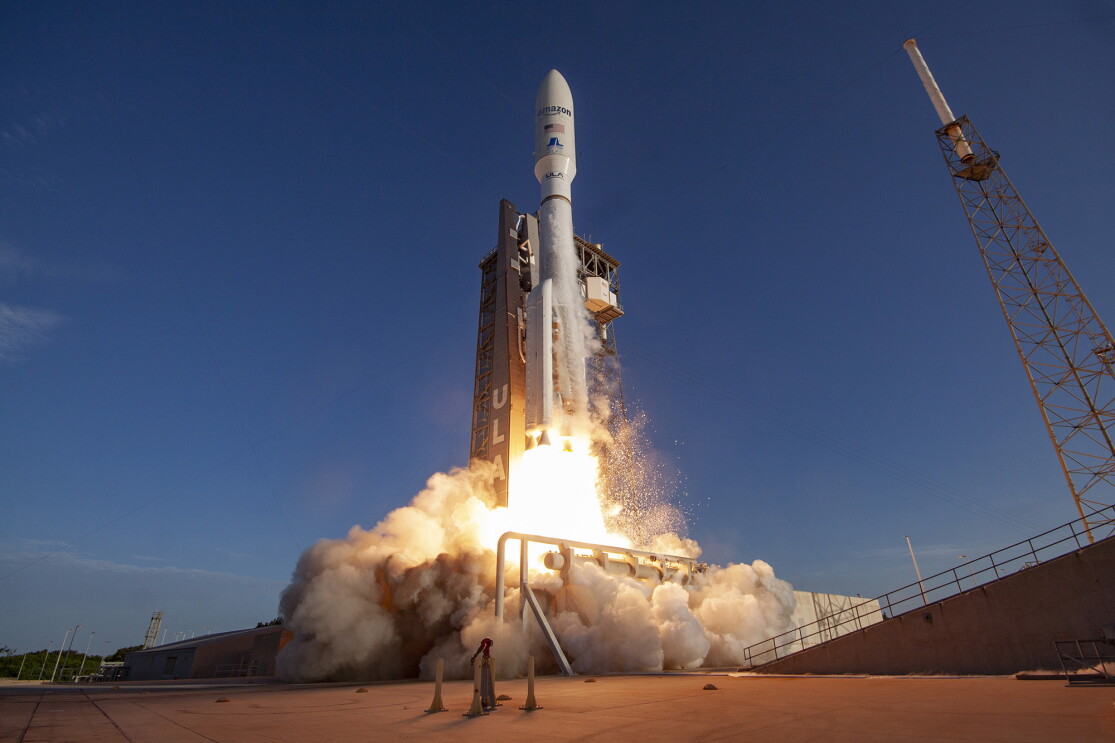 An image of a satellite launching. It has "Amazon" and the American flag decaled on the side. There is a blast below the satellite indicating launch and a blue, evening sky in the background.