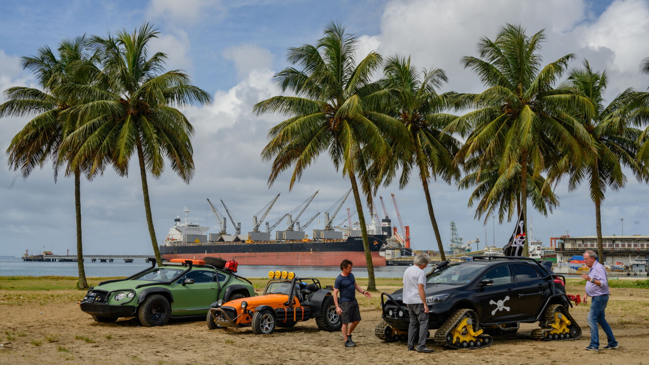 Three men stand next to three cars on a beach with palm trees in the background