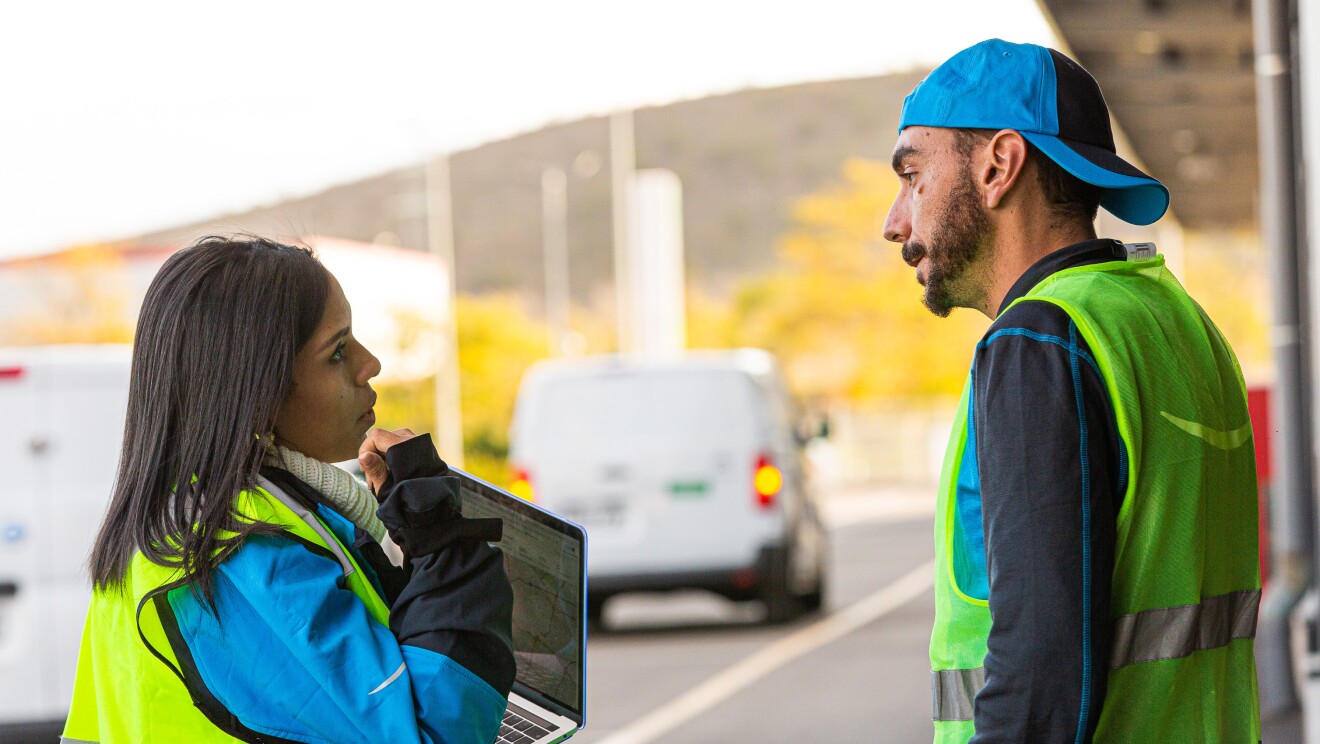 Ivette Duran, CEO de Cainaima Logistics, de pie con un ordenador entre las manos y hablando con un conductor de reparto de paquetes. Están los dos en la calle y de fondo hay dos furgonetas blancas. Ella es morena y tiene el pelo largo y negro. Lleva un jersey de color blanco y de cuello alto, una chaqueta azul y un chaleco fluorescente. Ell es más alto, con barba y bigote y lleva una chaqueta de color azul y gris y un chaleco fluorescente. Los dos están hablando cara a cara.