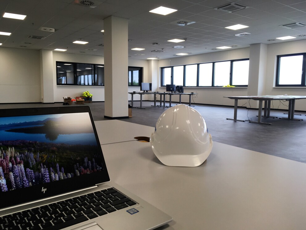 Auf einem weißen Schreibtisch steht links ein Laptop, rechts ein weißer Bauhelm. Im Hintergrund Schreibtische mit einigen Bildschirmen. Die Bürofläche ist noch nicht fertig eingerichtet.