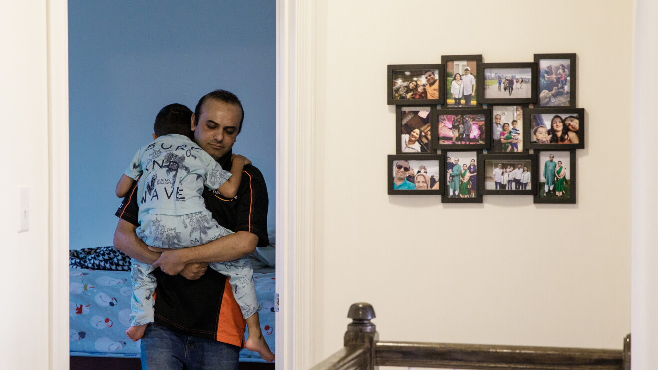 A man carrying a young boy steps out of the doorway of a bedroom. To the man's left are framed family photos. A staircase railing is visible in the foreground.
