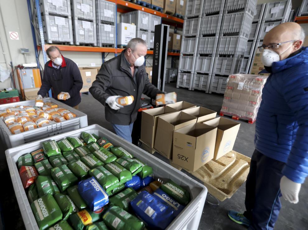 Banco de alimentos. En la parte inferior de la foto, dos cubetas grandes con paquetes de arroz y azúcar. Tres personas de mediana edad, con mascarilla, están con paquetes en las manos colocándolos en cajas de cartón. De fondo, estanterías llenas de palés de color gris. 