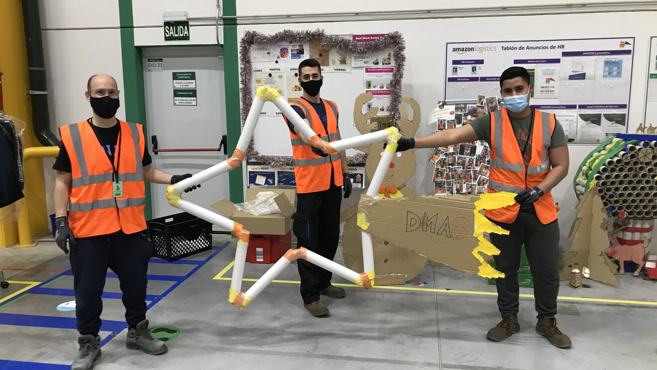 Adorno Navideño de DMA8. Tres trabajadores del centro están aguantando una estrella fugaz hecha de cartón y lleva el nombre del centro logístico. Ellos trs van vestidos con chalecos naranjas, pantalones negros y mascarillas. De fondo se ven dos tableros de trabajo. La foto está hecha dentro del centro logístico. 