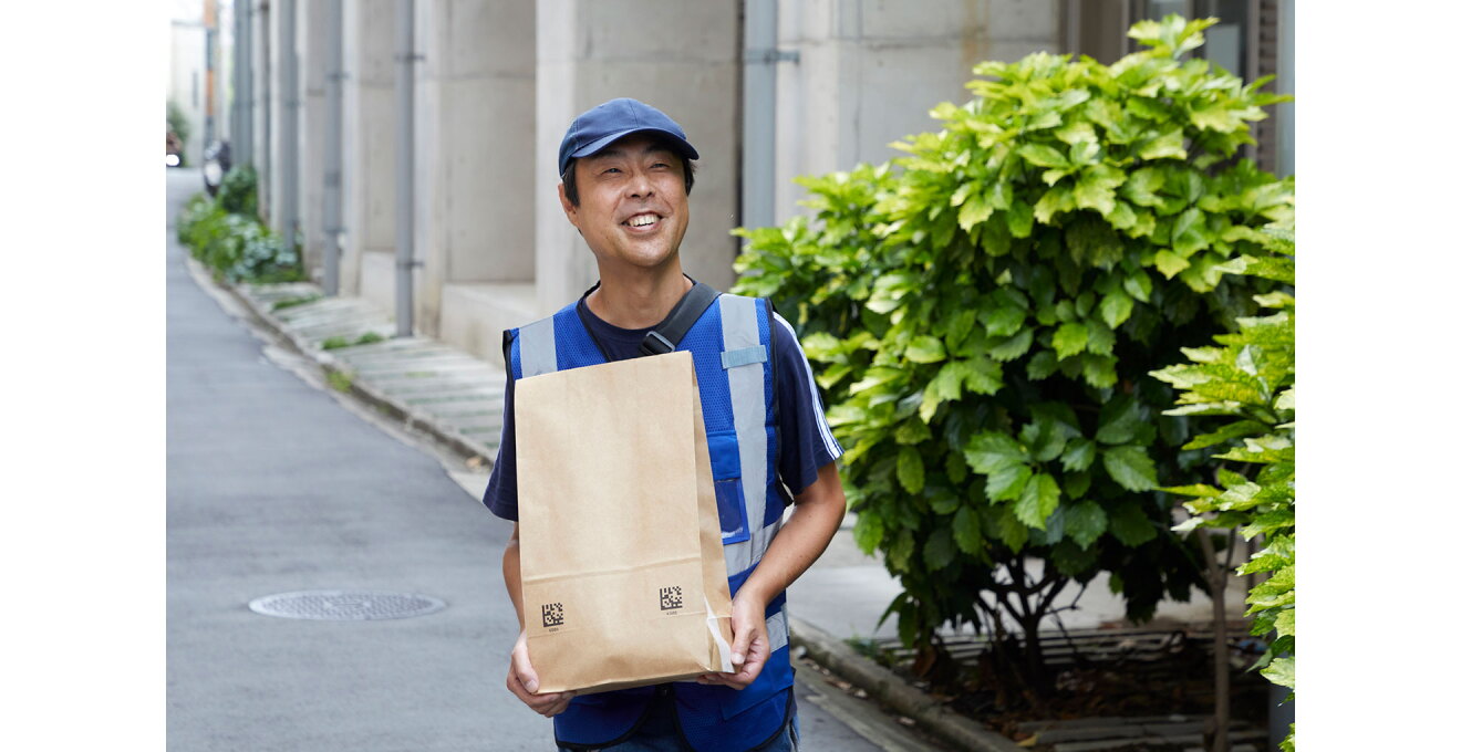 ガード下の道路を、クラフト用紙の紙袋を抱えて運ぶ配送スタッフ。キャップをかぶし、ベストを着用している。