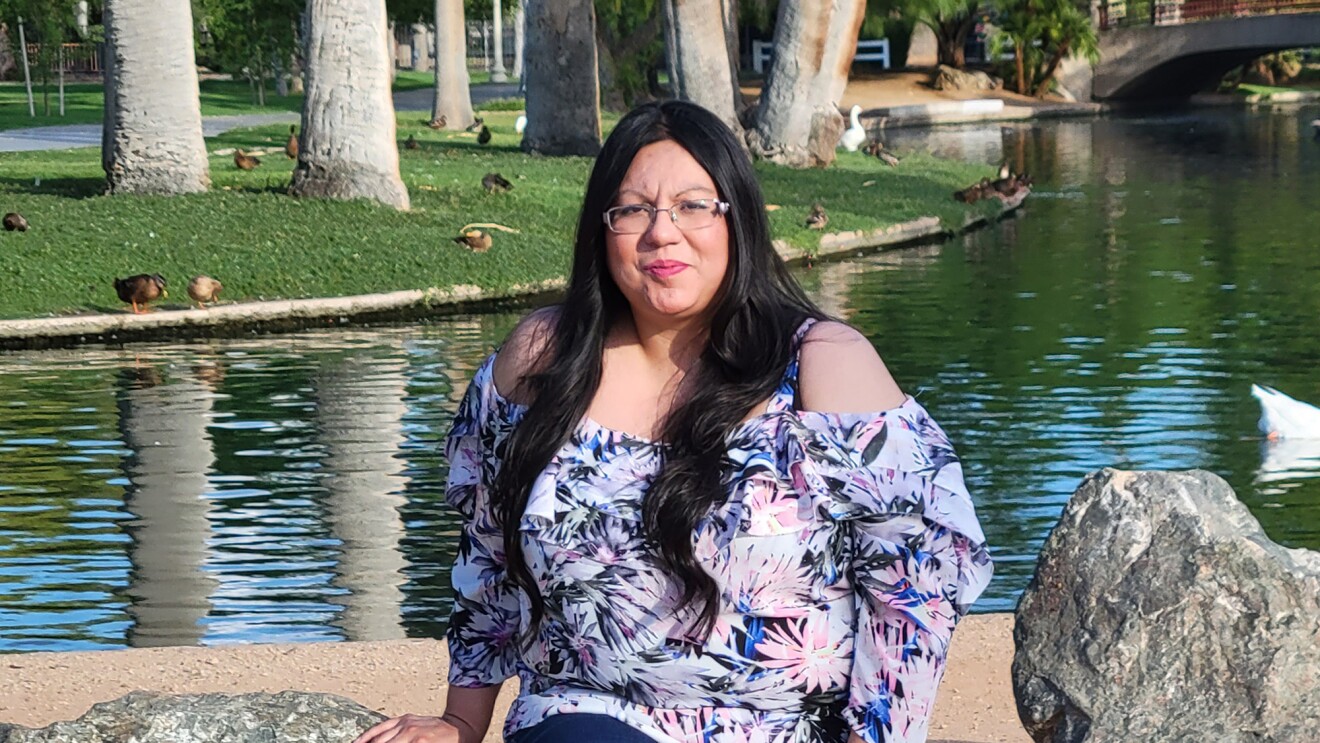 An image of a woman smiling for a photo while sitting on a bench in front of a pond at a park.