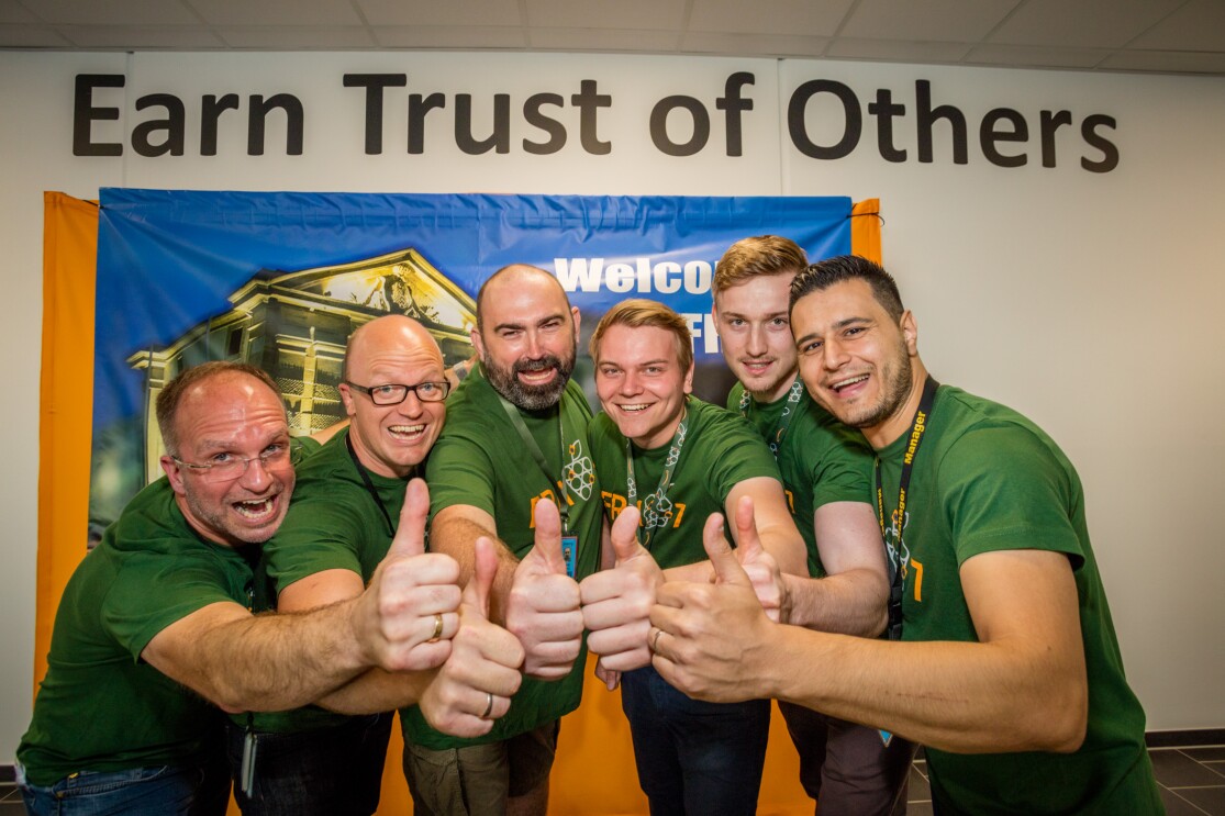 Sechs Mitarbeiter, männlich, alle mit grünen T-shirts und mit Daumen nach oben, stehen vor einem Willkommensplakat, das nicht vollständig sichtbar ist. Über dem Plakat eine Aufschrift: Earn trust for others. Alle Mitarbeiter lächeln.