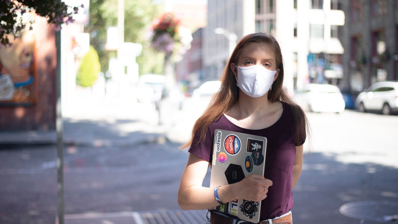 A woman stands on the street with a mask on, looking at the camera. She's holding a laptop with stickers related to Amazon and AWS.