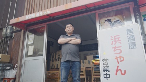 居酒屋の入り口で腕を組んで立つ男性。下から見上げる構図。引き戸が開いていて、店内が見える。