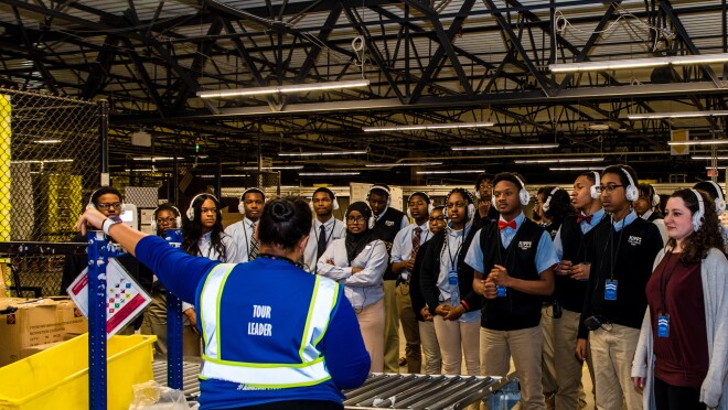 穿着校服、戴着耳机的高中生们站在亚马逊物流中心内。他们面对着一位亚马逊导游,她带着他们参观设施。