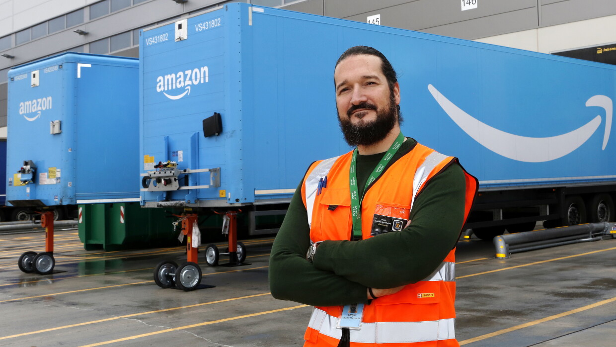 Sergio Amador con los brazos cruzados, un chaleco naranja, mira a cámara. Tiene barba y el pelo largo negro, recogido con una cola. De fondo están dos remolques azules con la sonrisa de Amazon. 