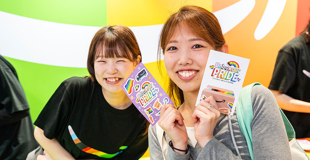 笑顔を見せる2人の女性。右側の女性は、両手に持った2枚のカードを見せている。左側の女性の黒いTシャツ前面にレインボーのロゴ。